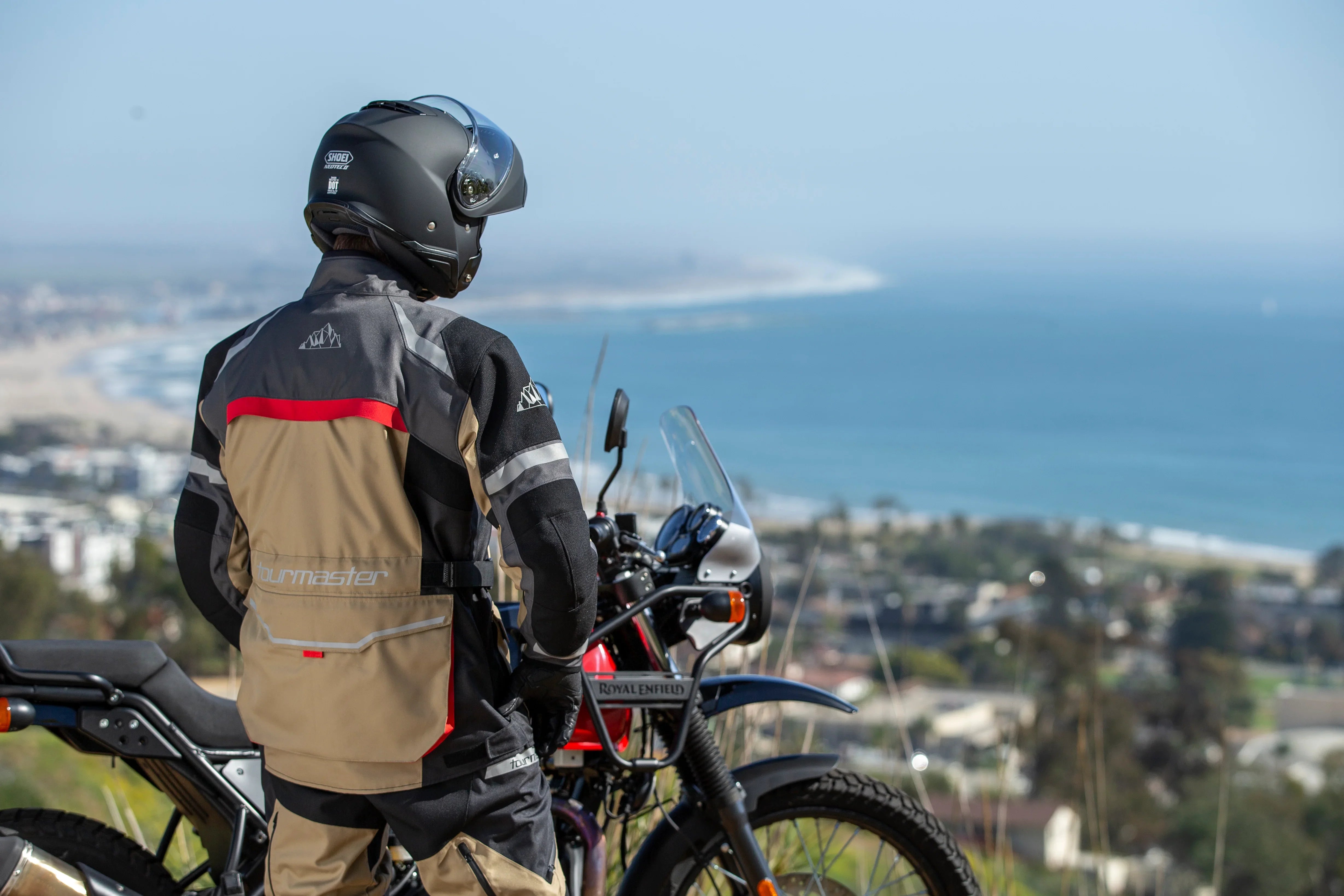 Motorcyclist wearing black helmet and Tourmaster jacket overlooking ocean with Royal Enfield bike